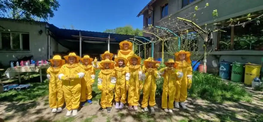 Група деца в жълти пчеларски костюми с шапки с мрежа стоят в редица в двор пред хижа.