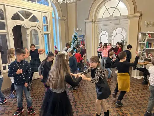 Пълна зала с танцуващи деца пред украсена елха и високи сводести прозорци.