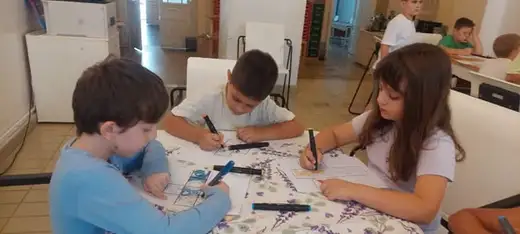 Три деца рисуват с флумастери, седнали край масата