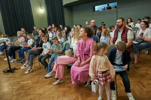 Зала с родители и деца, седнали в редици столове по време на церемонията.