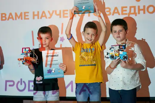 Три деца вдигат нагоре сертификати и роботи на сцена с надпис „Роботика“.