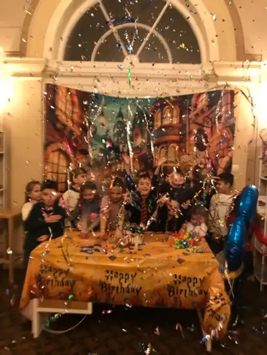 Деца стоят зад маса с покривка „Happy Birthday“, докато във въздуха се сипе конфети.